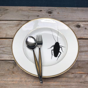 Puede incluir: Un plato de cerámica blanco con un borde dorado presenta una silueta negra de una cucaracha. Un tenedor y una cuchara de plata se colocan en el plato.