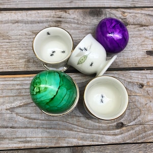 May include: Four egg cups with a gold rim, each with a different design. One cup has a green and white egg with a swirl pattern, another has a purple egg, and two cups have black ants on a white background.