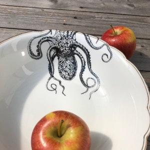 Peut inclure: Bol en céramique blanc avec une illustration de poulpe noir. Le bol a un bord festonné avec une bordure dorée. Deux pommes rouges sont à côté du bol.