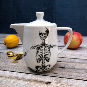 Op de afbeelding: Witte keramische theepot met een zwart skeletontwerp. De theepot heeft een deksel en een handvat.