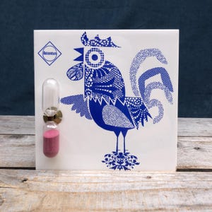 Puede incluir: Azulejo cuadrado blanco con un diseño de gallo azul y un pequeño reloj de arena. El gallo está decorado con patrones intrincados. El reloj de arena tiene un accesorio de latón y contiene arena rosa. El azulejo tiene la palabra "Boitzenburg" en un diamante azul.