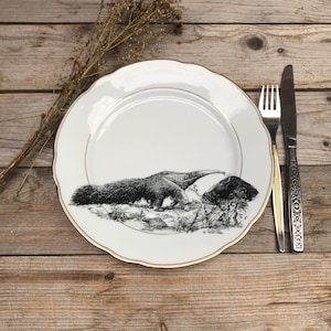 Op de afbeelding: Een wit keramiek dinerbord met een gouden rand heeft een zwart-witte illustratie van een miereneter. Het bord ligt op een houten oppervlak met een vork en mes ernaast.