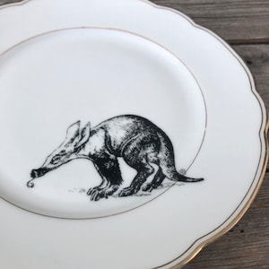 Pode incluir: Um prato de cerâmica branco com uma borda dourada apresenta uma ilustração em preto e branco de um oricteropo. O oricteropo é retratado em posição de caminhada, com seu longo focinho e cauda visíveis.