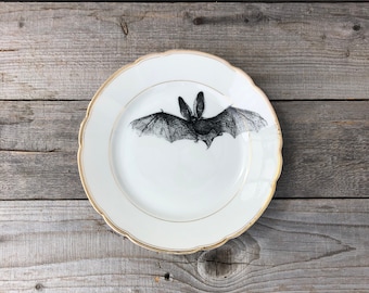 May include: A white ceramic plate with a gold rim features a black and white illustration of a bat.