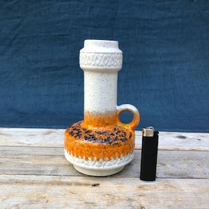 Puede incluir: Un jarrón de cerámica con una parte superior blanca moteada, un asa y una base con estampado naranja y negro. El jarrón tiene forma cilíndrica con un diseño texturizado alrededor de la parte superior e inferior. Un encendedor negro está al lado del jarrón.