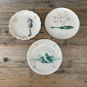 Pode incluir: Três pires de cerâmica branca com bordas douradas. Um apresenta uma ilustração de cavalo-marinho preto, outro mostra um peixe verde com detalhes florais rosa e verdes, e o terceiro exibe um gráfico de estrela-do-mar verde com pequenas estrelas coloridas.