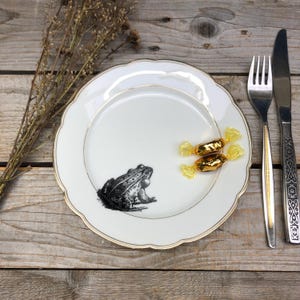 Può includere: Un piatto in porcellana bianca con un bordo dorato presenta un'illustrazione in bianco e nero di una rana. Due caramelle gialle sono sul piatto.