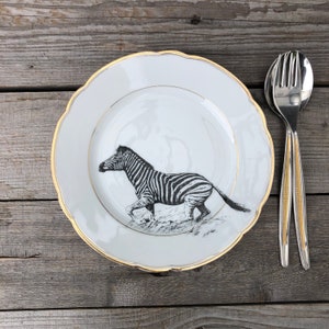 Könnte beinhalten: Weiße Keramikplatte mit einer schwarz-weißen Zebra-Illustration. Der Teller hat einen gewellten Rand mit Goldrand. Besteck mit goldenen Akzenten befindet sich rechts vom Teller. Auf einer Holzoberfläche.