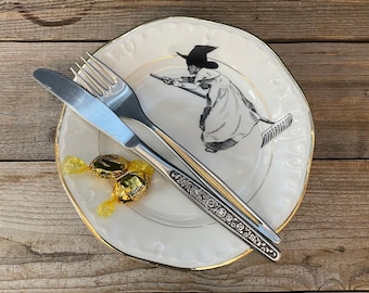 May include: A white plate with a gold rim featuring a black and white illustration of a witch on a broom. Silverware, including a fork and knife, is placed on the plate. Two wrapped candies are also on the plate.