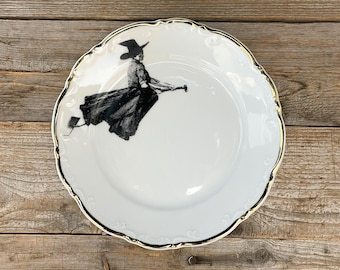 May include: A white ceramic plate with a scalloped edge and a gold-painted rim. The center features a black ink-style illustration of a figure wearing a pointed hat and a long skirt, riding a broomstick in a flying motion across the surface.