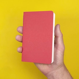 Puede incluir: Una libreta pequeña y rectangular con una cubierta de color rojo sólido y páginas blancas. La libreta se sostiene en posición vertical, mostrando su diseño simple y limpio y su color rojo vibrante sobre un fondo amarillo brillante.