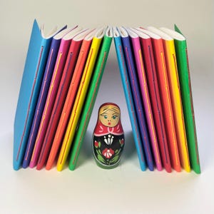 Può includere: Una collezione di dodici quaderni sottili e colorati disposti a forma di arco, con copertine blu, viola, rosa, arancioni, gialle e verdi e rilegature cucite a vista. Una bambola russa in legno dipinta si trova al di sotto.