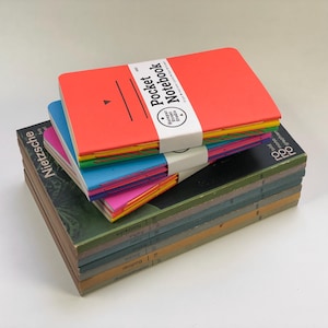 May include: A stack of books topped with three colourful pocket notebooks. The top notebook is bright orange with the text Pocket Notebook and Hotspur Street Studio. The notebooks have exposed stitching in yellow, blue, and pink, resting on vintage books.