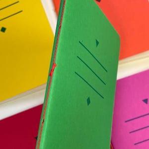 May include: A bright green notebook with a red stitched spine. The cover features a dark green graphic design consisting of three horizontal lines flanked by a small triangle at the top and a diamond shape at the bottom, centred on the front cover.