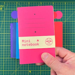 May include: A selection of mini notebooks in shades of pink, purple, blue, and red. The front notebook is a bright pink with the text "Mini notebook" and the Hotspot Street logo. The notebooks are held by a hand.