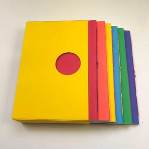 Può includere: Un set di quaderni rettangolari con un cofanetto esterno giallo dotato di un'apertura circolare. I quaderni sono disposti in fila, mostrando uno spettro di colori che include copertine rosse, arancioni, gialle, blu, verdi e viola con cuciture a vista.