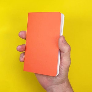 May include: A small, rectangular notebook with a solid, vibrant orange cover. The notebook features a clean, minimalist design with white pages visible along the side edge, providing a sharp contrast against the bright orange front cover.