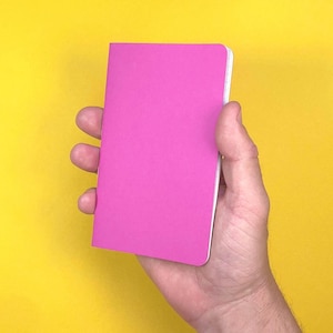 Puede incluir: Una libreta pequeña y rectangular con una cubierta de color rosa intenso sólido. La libreta presenta un diseño limpio con esquinas redondeadas y un borde blanco visible en el lomo, presentada en orientación vertical sobre un fondo amarillo.