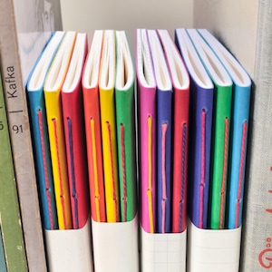 Op de afbeelding: Een verzameling van tien slanke notitieboekjes met felgekleurde kartonnen omslagen in blauw, geel, rood, groen, roze en paars. Elk boekje is voorzien van een zichtbare, met de hand genaaide binding met contrasterend garen en wit papier.