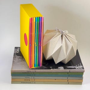 Op de afbeelding: Een felgele cassette met vijf dunne, kleurrijke boekjes met zichtbare stiksels, naast een beige, geometrisch gevouwen papieren sculptuur. Deze items staan op een stapel van vijf boeken met neutrale ruggen waarop de namen Kafka, Nietzsche, Keller, Sartre, Büchner en Hofmannsthal staan.