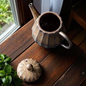 以下が含まれることがあります： 濃い茶色と金色のセラミックティーポット。リブ模様と別蓋が特徴です。ティーポットは木製の表面に置かれています。注ぎ口とハンドルが見え、お茶を淹れるための機能的なデザインを示唆しています。