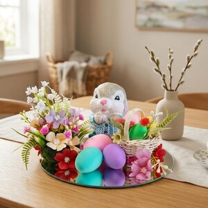 Könnte beinhalten: Eine Osterdekoration mit einer Keramik-Hasenfigur, pastellfarbenen Eiern und einem kleinen Weidenkorb mit Eiern und Blumen. Die Anordnung umfasst eine Spiegelbasis und eine Vase mit Kätzchenzweigen.