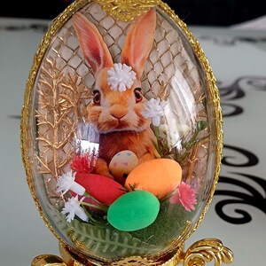 May include: An Easter-themed decorative egg with a gold filigree frame and stand. Inside, a bunny with a flower crown sits among colorful eggs and faux greenery. The egg is approximately 6 inches tall.