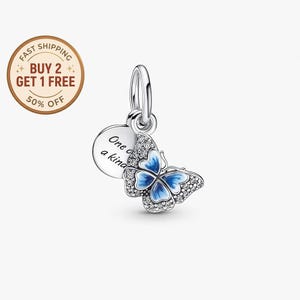 Puede incluir: Un charm de plata con un diseño de mariposa. La mariposa tiene alas de esmalte azul y está adornada con pequeños cristales transparentes. Un charm redondo con el texto "One of a kind" cuelga junto a la mariposa. El charm está diseñado para colgar de una pulsera o collar.