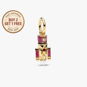May include: A gold-colored charm in the shape of a nutcracker soldier. The charm has a pink glittery hat with a star and a clear gem. The soldier has white boots and pink accents. The charm is a festive holiday item.