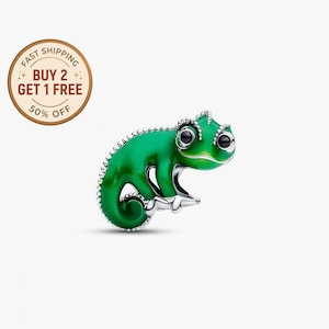 Puede incluir: Un charm de camaleón verde con ojos negros y detalles plateados. El charm tiene un acabado brillante y está en posición sentada. Es una pieza de joyería.
