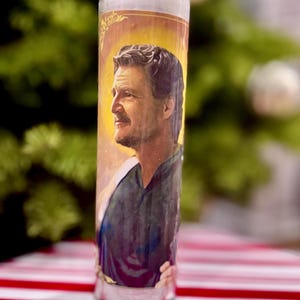 May include: A clear glass candle with a portrait of a man on the front. The portrait is set against a gold background with a halo. The candle sits on a red and white striped surface.