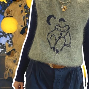 Peut inclure: Un gilet en tricot vert sauge avec un motif sombre d'une figure. Superposé à un haut à manches longues bleu marine et associé à un pantalon plissé bleu marine. Un collier doré et une ceinture marron complètent le look.