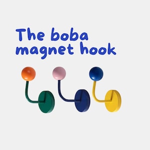Könnte beinhalten: Ein Set aus drei farbenfrohen Magnet-Haken. Jeder Haken hat eine runde, farbige Kugel auf einem gebogenen Metallarm. Der Text "The boba magnet hook" steht in Blau über den Haken. Die Haken sind orange, rosa und blau.