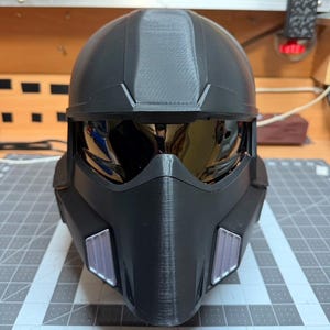 Könnte beinhalten: Ein schwarzer Helm mit Visier und strukturierter Oberfläche. Der Helm hat eine abgerundete Oberseite und kantige Merkmale, mit einem reflektierenden Visier und Akzenten an den Seiten. Das Design deutet auf ein futuristisches oder Science-Fiction-Thema hin.