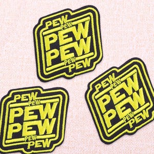 Puede incluir: Tres parches bordados con la palabra "PEW" en un patrón repetido. Los parches tienen un borde negro y el texto es de color amarillo brillante. Los parches están sobre una tela beige claro.