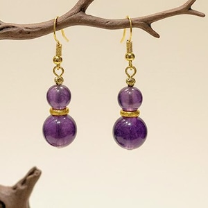 May include: A pair of dangle earrings featuring two purple, spherical beads connected by a gold-colored metal. The earrings hang from gold-colored ear wires. The earrings are displayed on a brown branch against a neutral background.