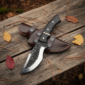 May include: A handcrafted hunting knife with a black handle and a textured blade, resting in a brown leather sheath. The knife is displayed on a rustic wooden surface with fallen leaves.