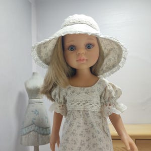 Peut inclure: Une poupée portant un chapeau de soleil crème avec un motif floral et une bordure en dentelle. La poupée a les cheveux blonds et porte une robe avec un motif floral et des détails en dentelle. La poupée est devant un fond blanc.