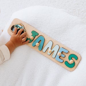 May include: Wooden name puzzle spelling "JAMES" in teal, light blue, and green letters. The puzzle includes a truck graphic and wooden pegs for easy handling. The puzzle is made of light-colored wood.