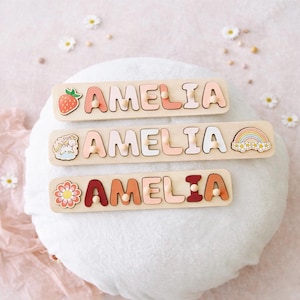 K&ouml;nnte beinhalten: Drei Holz-Namenspuzzles mit dem Namen AMELIA in verschiedenen Rosa-, Orange- und Rott&ouml;nen. Jedes Puzzle hat einen kleinen Holzknopf an jedem Buchstaben sowie dekorative Symbole wie eine Erdbeere, ein Einhorn und eine Blume.