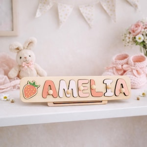 K&ouml;nnte beinhalten: Ein rechteckiges Namenspuzzle aus Holz mit dem Namen AMELIA in abwechselnd hellrosa und wei&szlig;en Gro&szlig;buchstaben sowie einem Erdbeersymbol auf der linken Seite. Jeder Buchstabe ist mit einem kleinen Holzknopf zum einfachen Greifen versehen.
