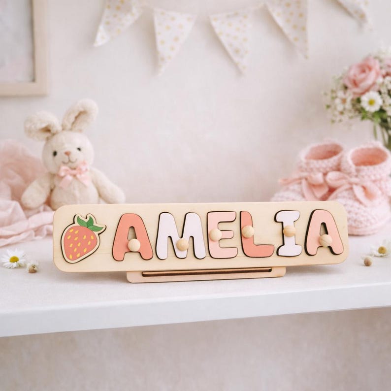 K&ouml;nnte beinhalten: Ein rechteckiges Namenspuzzle aus Holz mit dem Namen AMELIA in abwechselnd hellrosa und wei&szlig;en Gro&szlig;buchstaben sowie einem Erdbeersymbol auf der linken Seite. Jeder Buchstabe ist mit einem kleinen Holzknopf zum einfachen Greifen versehen.