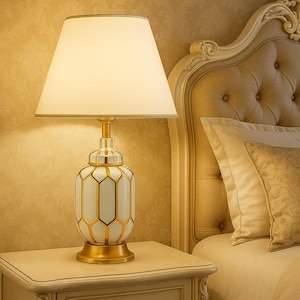 Puede incluir: Una lámpara de mesa blanca y dorada con un diseño geométrico, rematada con una pantalla de color crema. La lámpara está sobre una mesita de noche junto a una cama con cabecero acolchado y almohadas decorativas. La base de la lámpara mide unos 20 cm de diámetro.