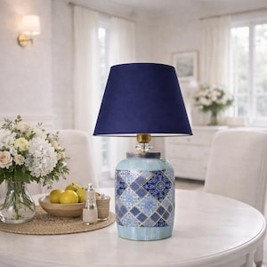 Puede incluir: Lámpara de mesa decorativa con pantalla azul marino. La base de cerámica presenta un patrón geométrico azul y blanco. La lámpara está sobre una mesa blanca en una habitación luminosa. Un elemento decorativo para el hogar.