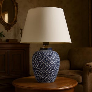 Może przedstawiać: Granatowa ceramiczna lampa stołowa z kremowym abażurem. Podstawa lampy ma biały geometryczny wzór. Lampa stoi na okrągłym drewnianym stole, tworząc ciepłą atmosferę. Wysokość około 60 cm.