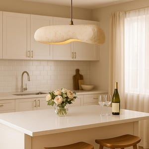 Puede incluir: Una cocina moderna con una lámpara colgante oblonga y única, con acabado de latón. La isla de la cocina tiene un ramo de rosas, copas de vino y una botella de vino. Hay dos taburetes de madera.