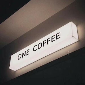 Op de afbeelding: Een rechthoekig, verlicht bord met de tekst "ONE COFFEE" in zwarte letters. Het bord is wit en gemonteerd op een donkere ondergrond, waarschijnlijk een muur of plafond. De algehele esthetiek is minimalistisch en modern.