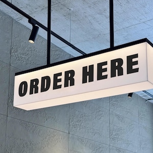 Op de afbeelding: Een rechthoekig, verlicht bord met de tekst "ORDER HERE" in vette, zwarte hoofdletters. Het bord is met zwarte metalen steunen aan het plafond bevestigd. De achtergrond is een getextureerde, grijze muur.