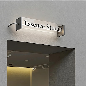 Puede incluir: Un letrero rectangular iluminado con el texto "Essence Studio" en letras negras. El letrero está montado en una pared gris sobre una puerta. Tiene una parte frontal transparente y un marco gris oscuro.