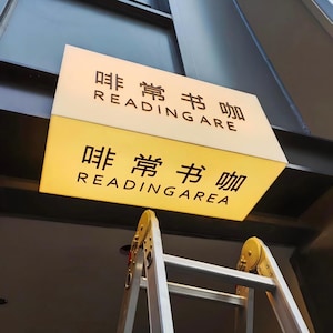 Puede incluir: Letrero rectangular iluminado con el texto "非常书咖 READING AREA" en negro. El letrero tiene una parte superior blanca y una inferior amarilla. Una escalera metálica está debajo.